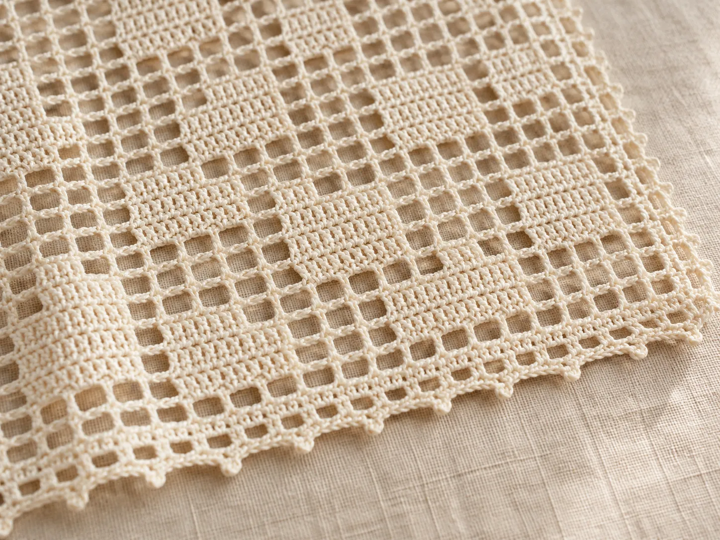 Close-up of ivory filet crochet lace with open mesh windows, filled geometric blocks, and a looped edging on linen